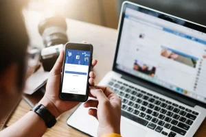 Facebook posztolás telefonról, háttérben egy laptop, amelyen a Facebook oldala van megnyitva